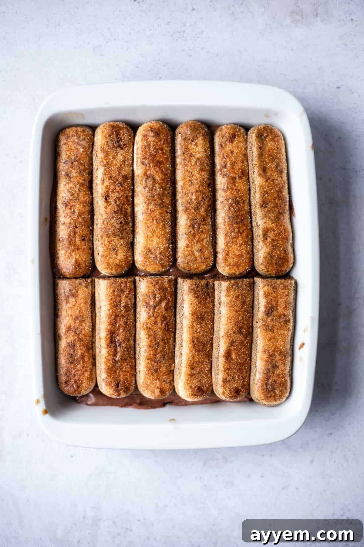 A baking dish displaying multiple layers of coffee-soaked ladyfingers alternating with rich chocolate mascarpone cream, ready for chilling.