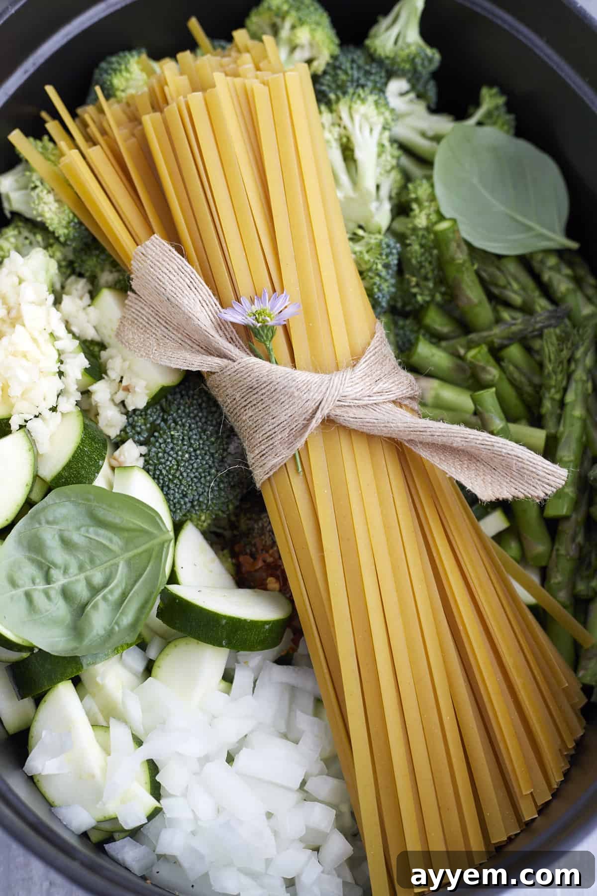 Overhead image of a bundle of pasta noodles over onions, zucchini, garlic, herbs, broccoli, and asparagus, ready to be cooked together.