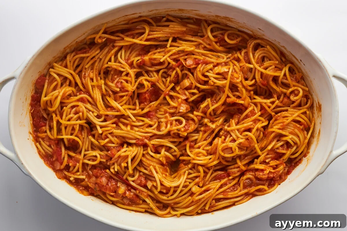 Baked spaghetti in an oval baking dish, halfway through cooking, showing the noodles absorbing the sauce.