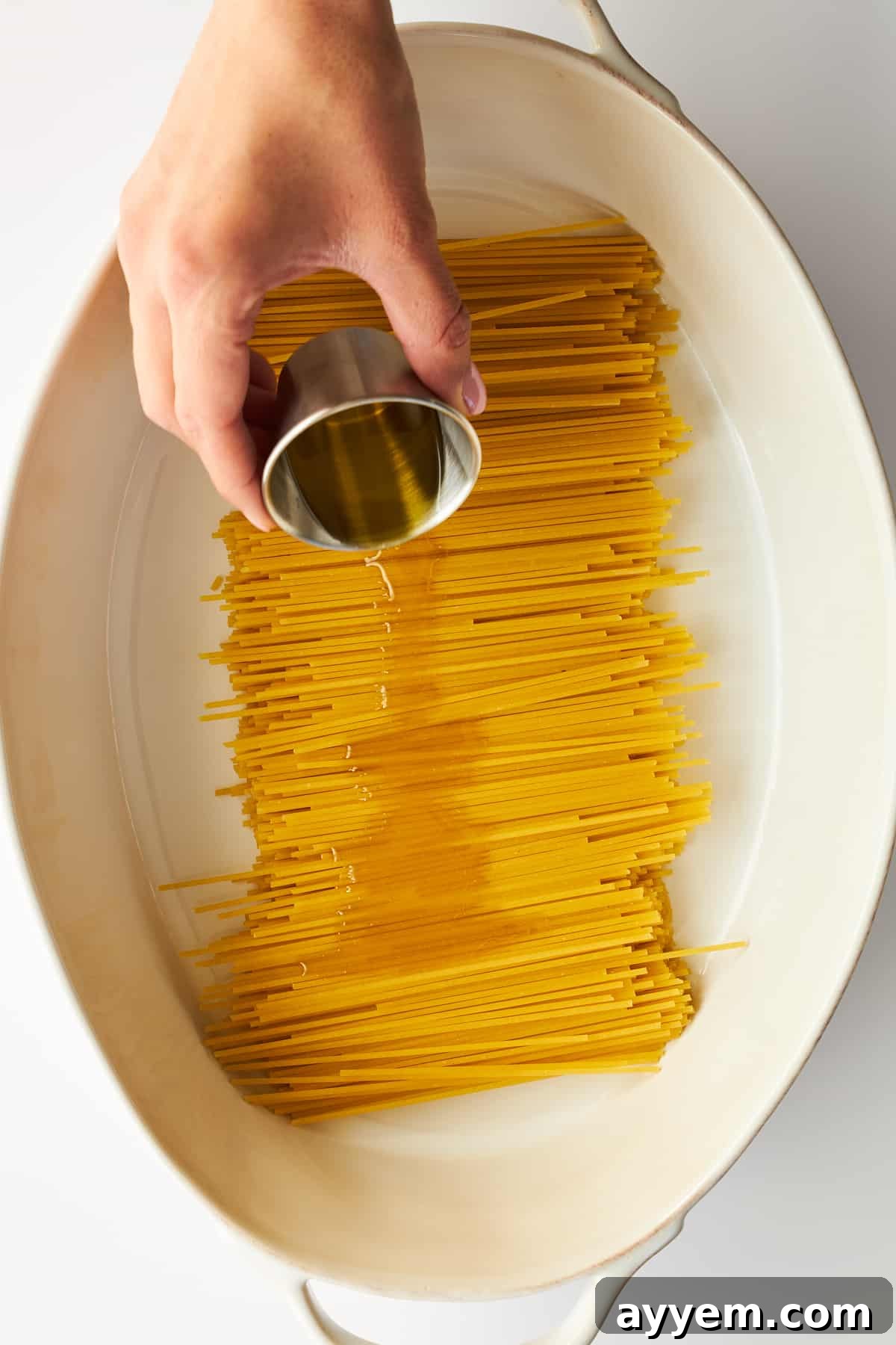Olive oil being drizzled generously over a bed of uncooked spaghetti noodles in an oval baking dish, preparing them for an easy oven-baked meal.