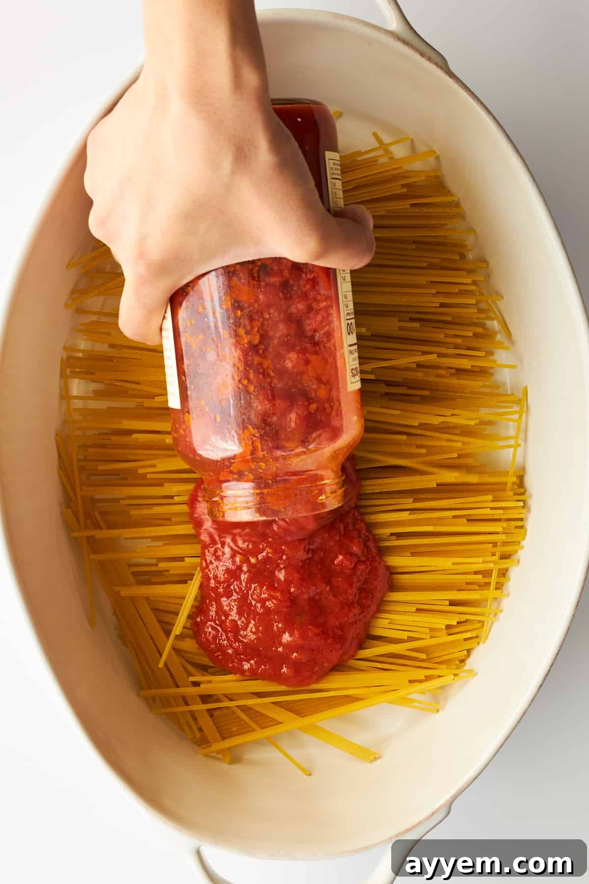 Rich marinara sauce being poured evenly over uncooked spaghetti noodles, ensuring every strand is coated for a flavorful oven-baked dish.