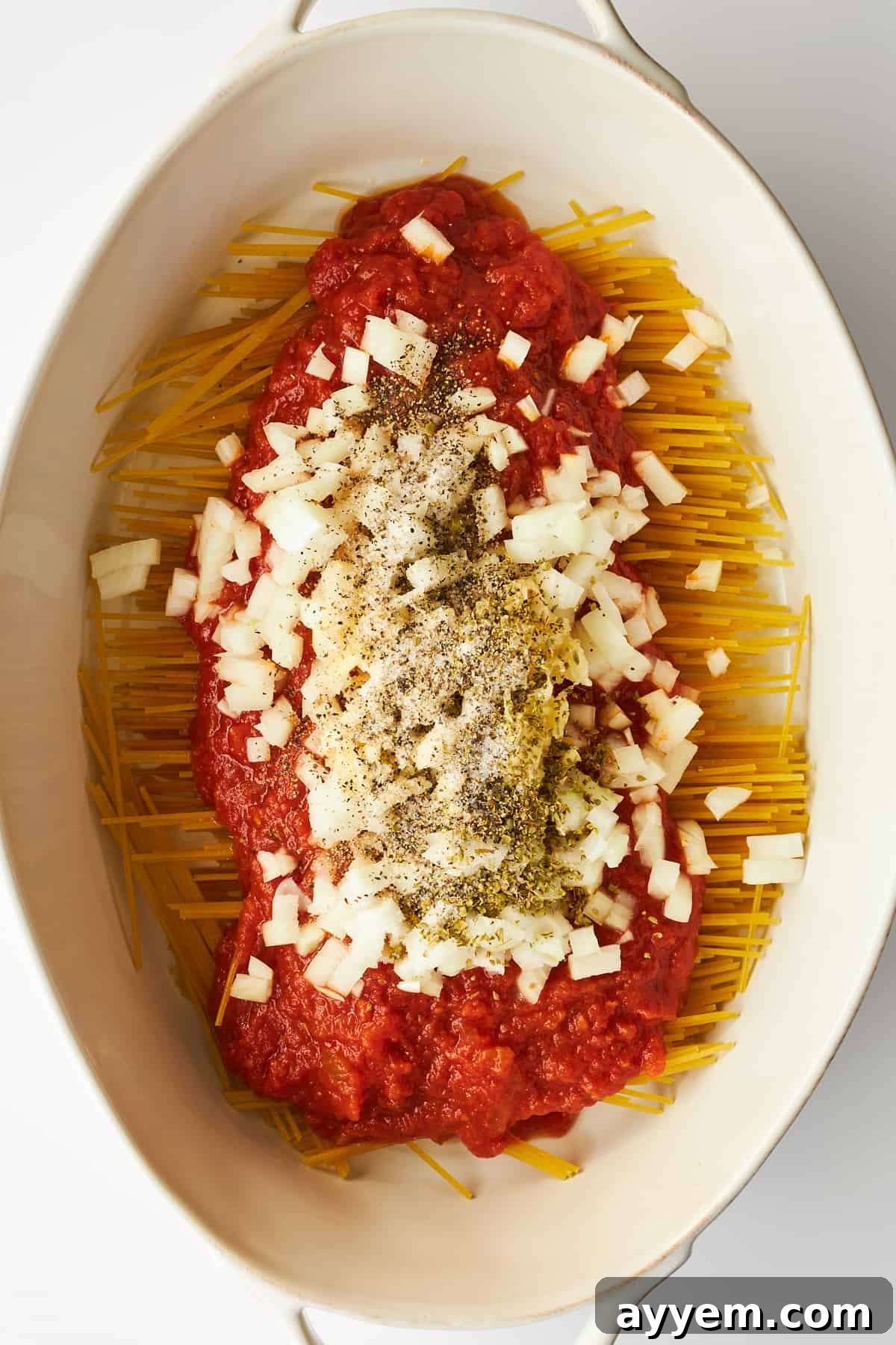 Finely chopped onions and aromatic seasonings, including oregano and black pepper, scattered over a layer of marinara-covered spaghetti in a baking dish, ready for mixing and baking.