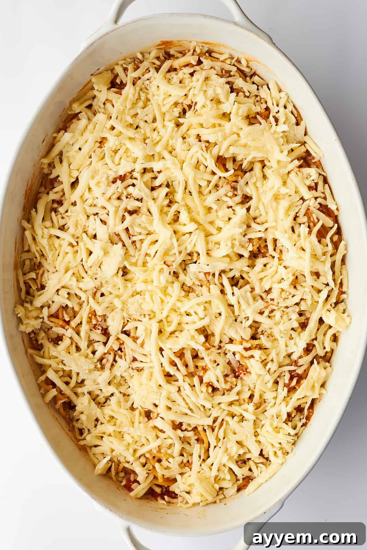 A generous layer of shredded mozzarella cheese sprinkled over the hot, baked spaghetti in an oval dish, anticipating its final bake to become golden and bubbly.