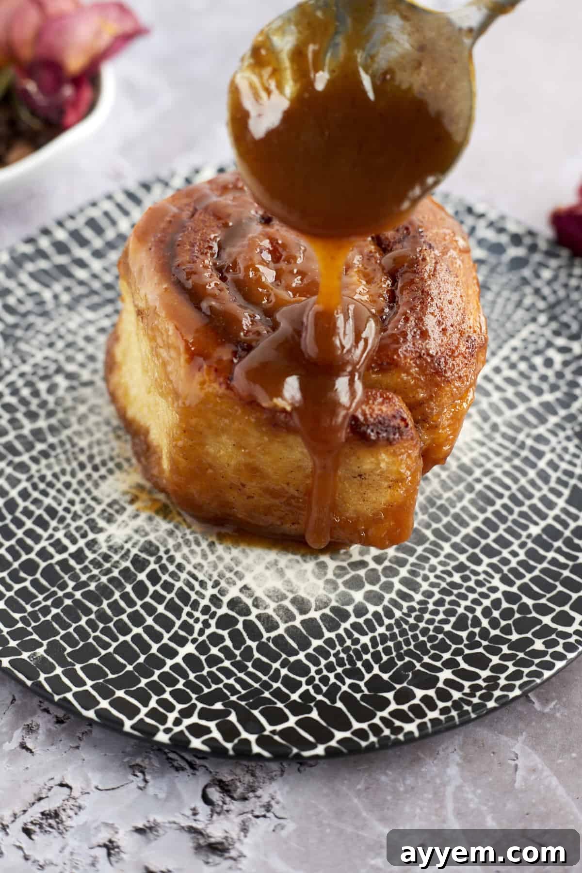 Caramel sauce being generously drizzled over a warm caramel macchiato cinnamon roll on a plate, ready to be enjoyed.