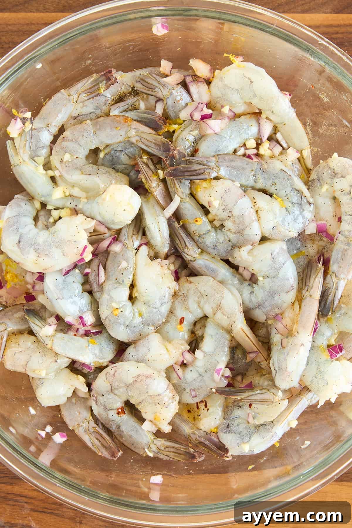 Raw shrimp marinating in a glass bowl with chopped red onions, minced garlic, lemon zest, and spices on a wooden surface.