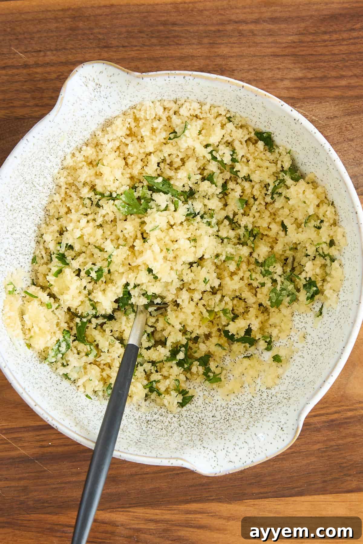 A white speckled bowl filled with a mixture of breadcrumbs, chopped herbs, and grated cheese sits on a wooden surface. A spoon rests inside the bowl.