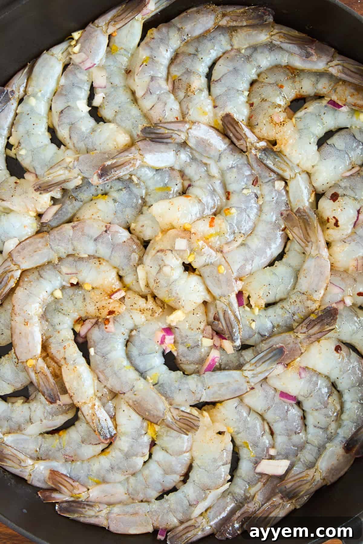 A close-up of raw shrimp arranged in a circular pattern in a pan, sprinkled with chopped garlic, red onion, and spices.