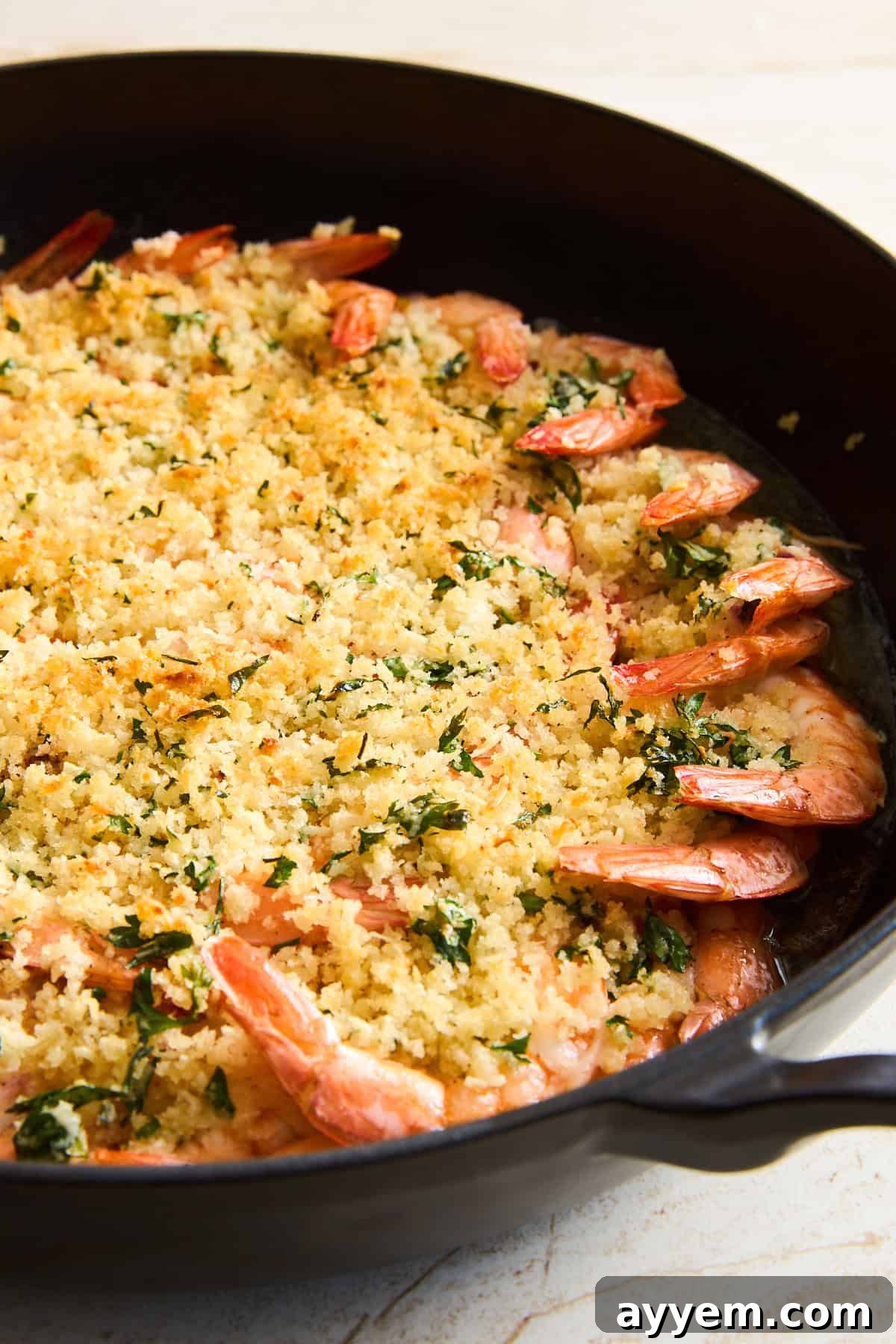 Shrimp baked in a black skillet, topped with golden, crispy breadcrumbs mixed with herbs. The shrimp are arranged in a circular pattern along the edge of the pan.
