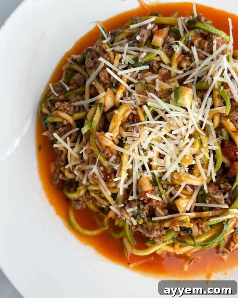 A serving of Low Carb Baked Spaghetti Zoodles in a white bowl, generously topped with fresh Parmesan cheese and a sprig of basil.