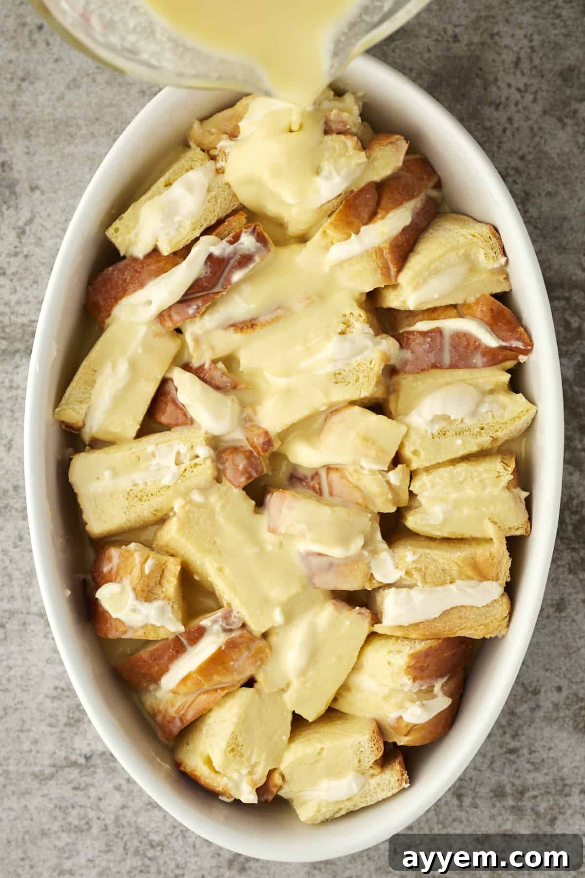 Ricotta Cream French Toast Bake 4 A hand pouring rich egg custard over unbaked cannoli french toast arranged in a baking dish.