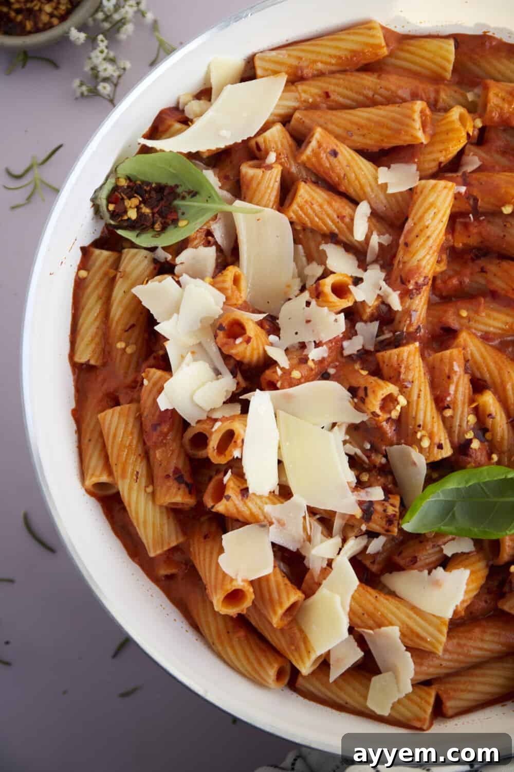 A large serving bowl filled with spicy rigatoni pasta, beautifully garnished with fresh shaved Parmesan cheese and a sprinkle of vibrant red pepper flakes.