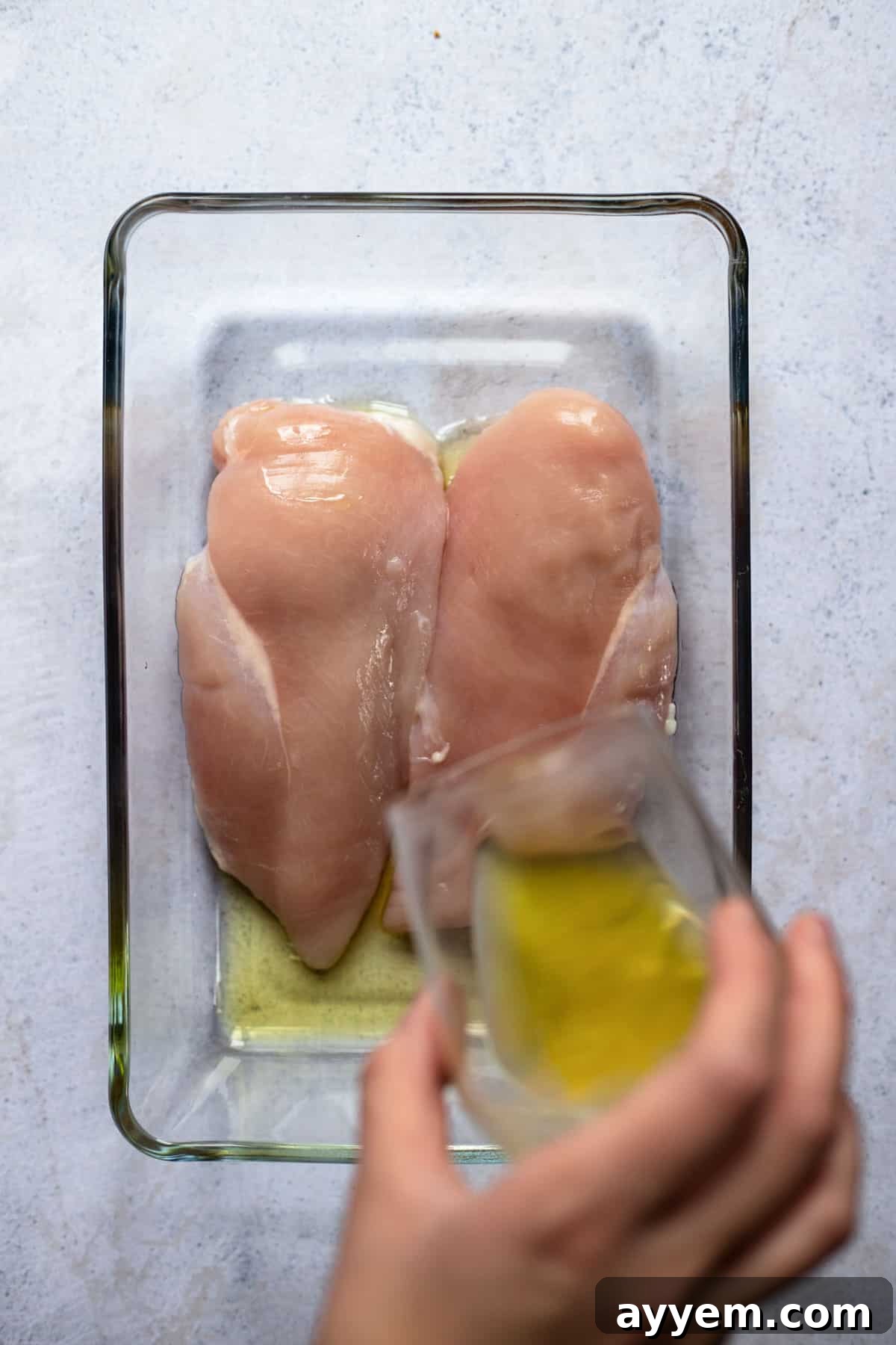 two raw chicken breasts in a baking dish with oil being poured over them 