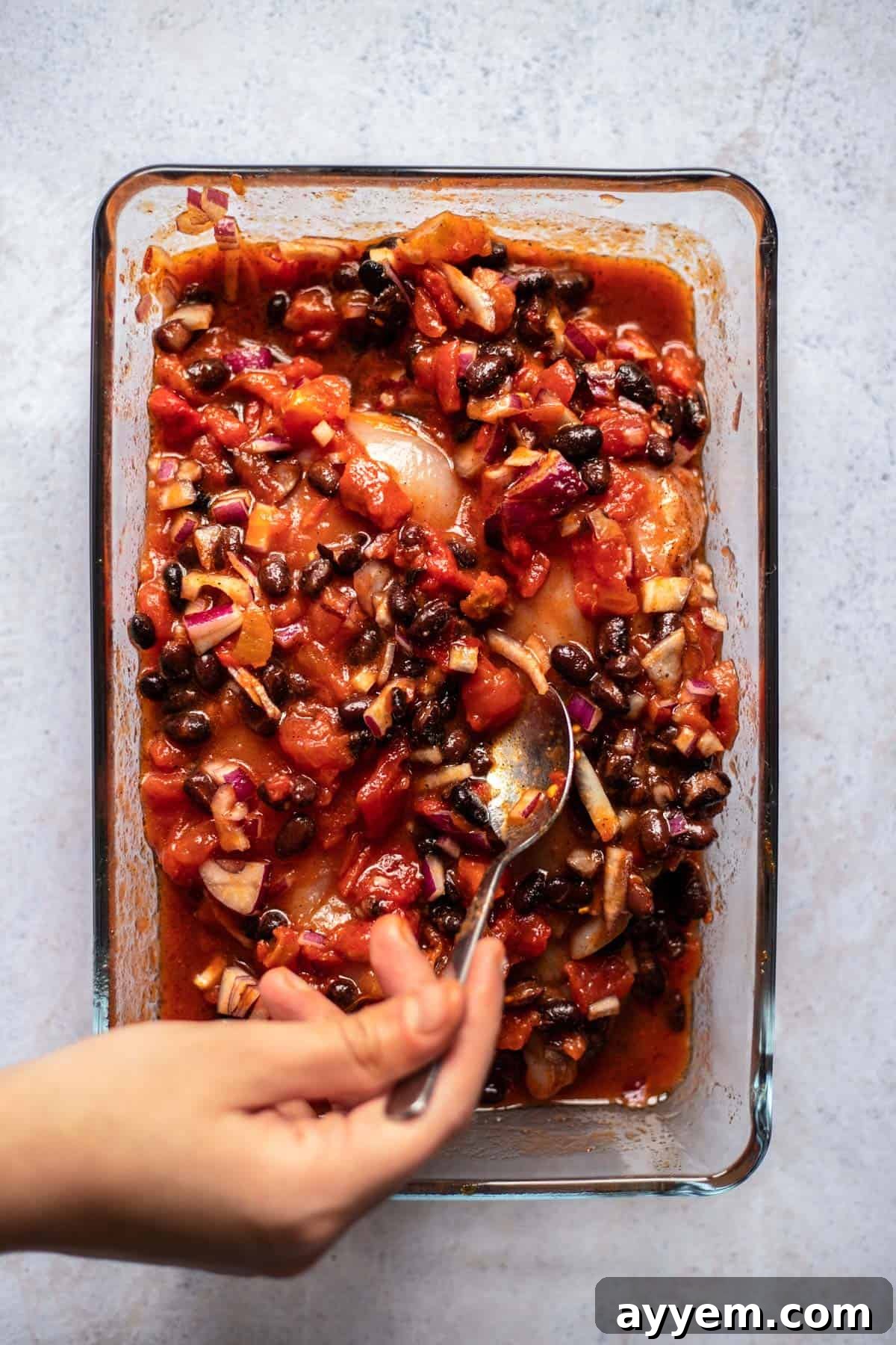 a hand using a spoon to stir salsa, beans, and red onions over two raw chicken breasts