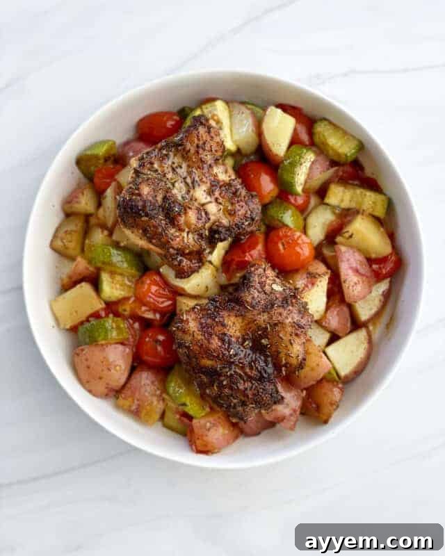 overhead image of a white bowl full of Sheet Pan Chicken Thighs and Veggies, garnished with fresh herbs.