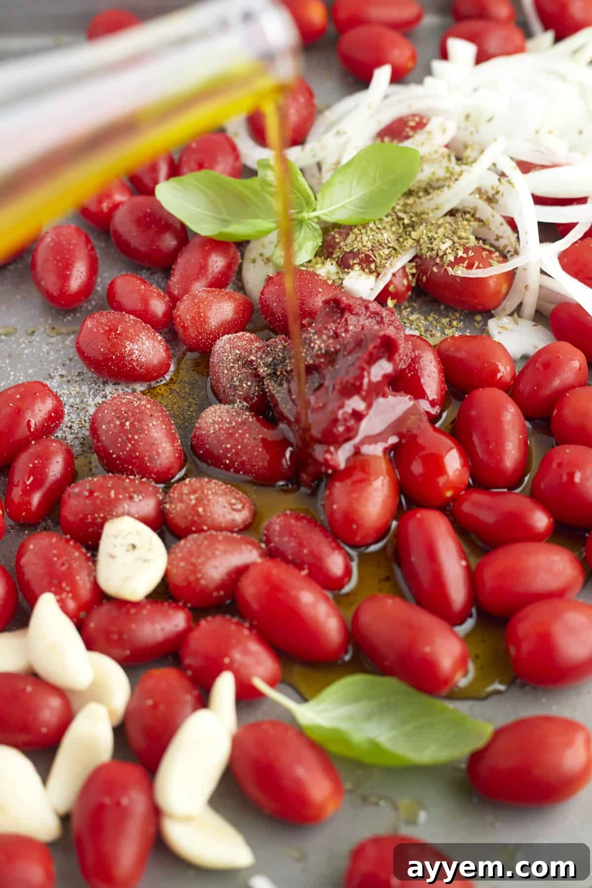 garlic cloves, tomatoes, and onions on a baking sheet topped with seasonings with olive oil being drizzled on top