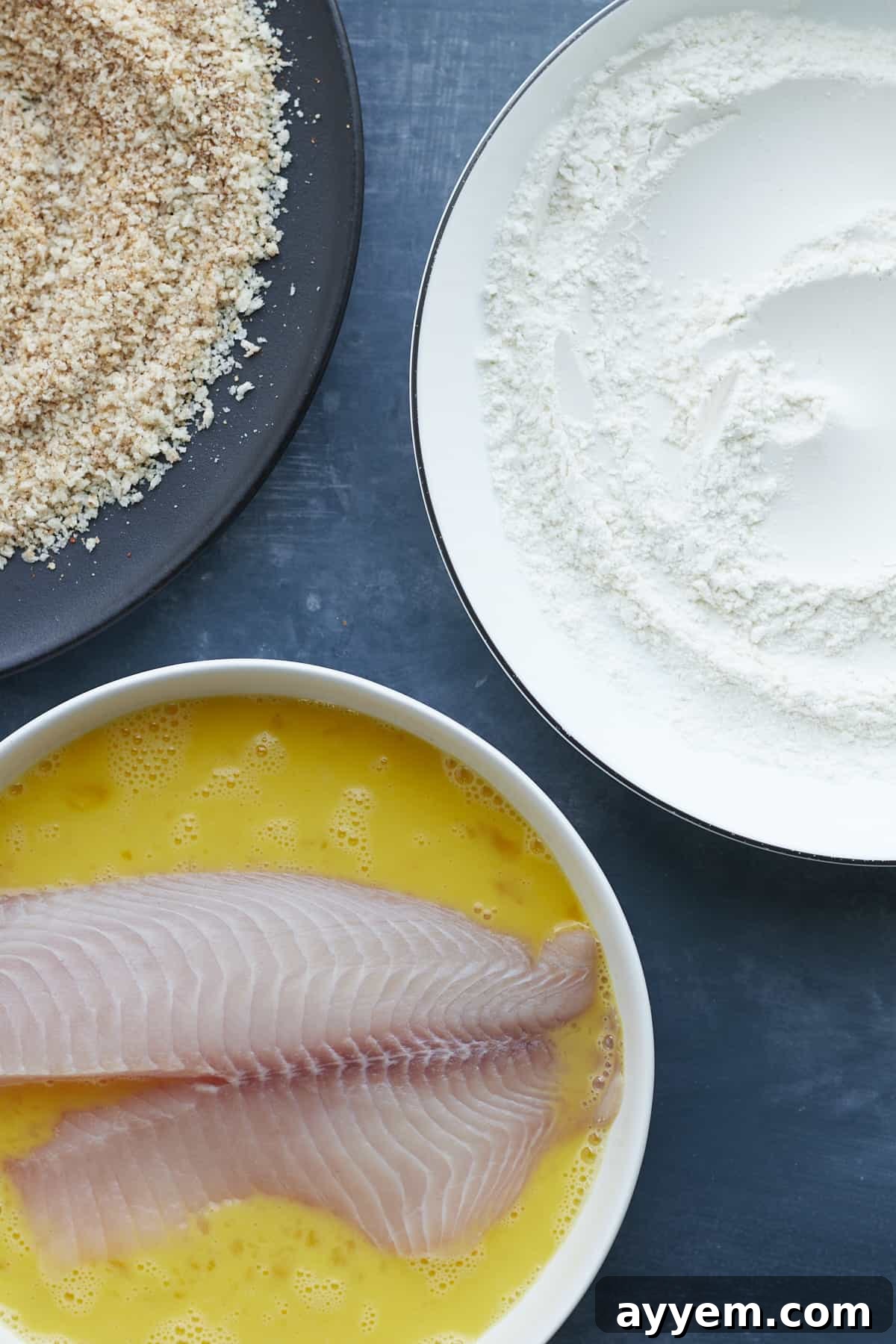 Tilapia fillet being dredged in egg wash, with bowls of flour and breadcrumbs to the side.