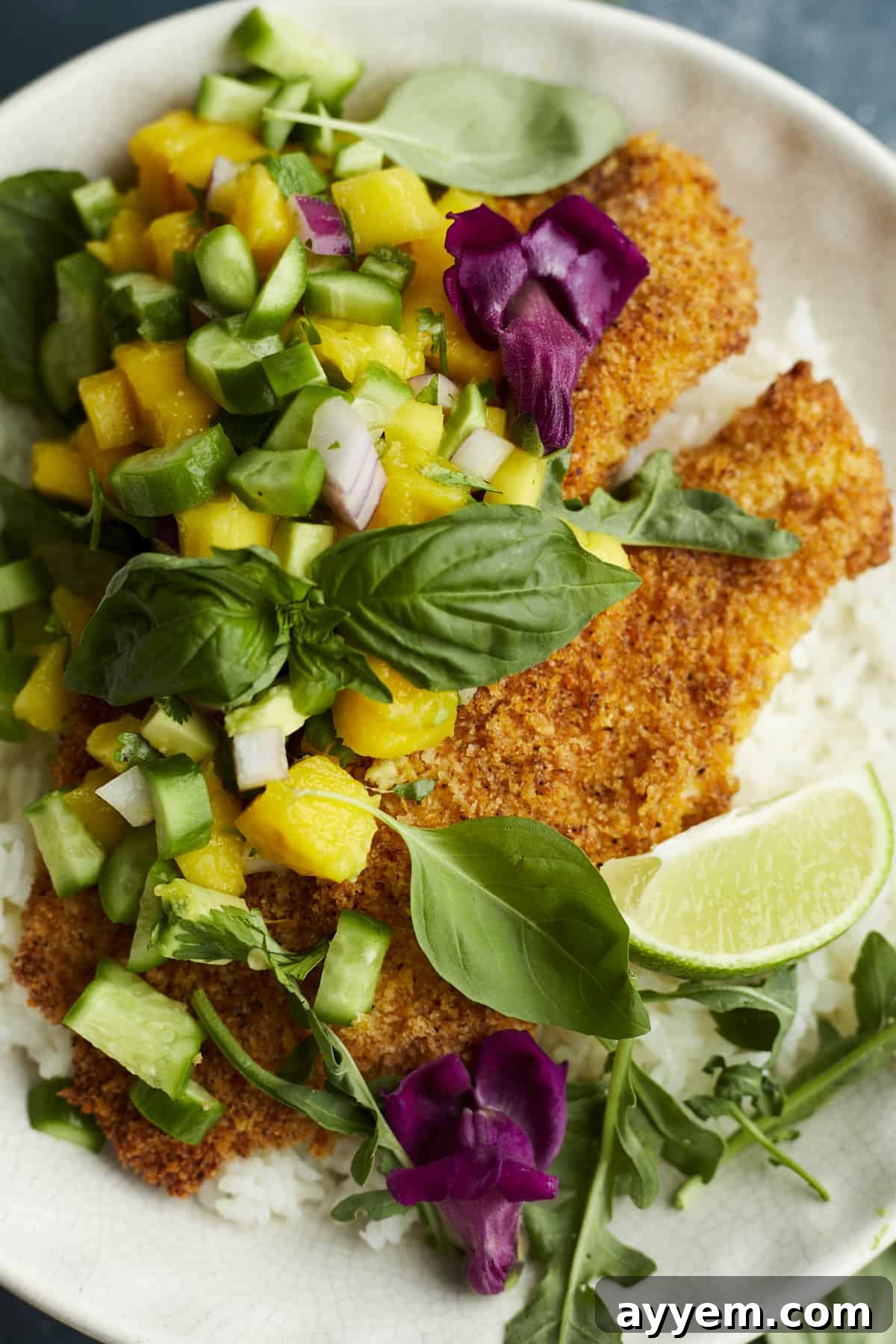 A white plate with a bed of fluffy coconut rice, topped with golden air fried tilapia and a generous scoop of fresh mango salsa, garnished with a lime wedge and cilantro.