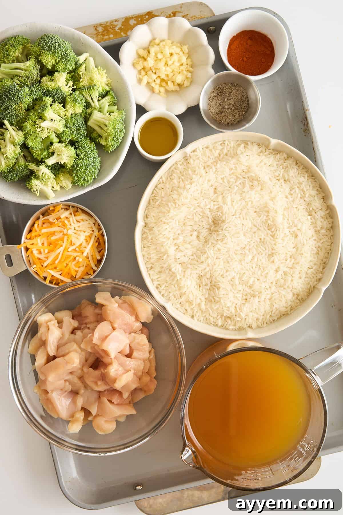 An overhead view of a tray holding bowls of raw broccoli, chopped garlic, shredded cheese, uncooked rice, diced raw chicken, chicken broth, olive oil, and small dishes of spices.