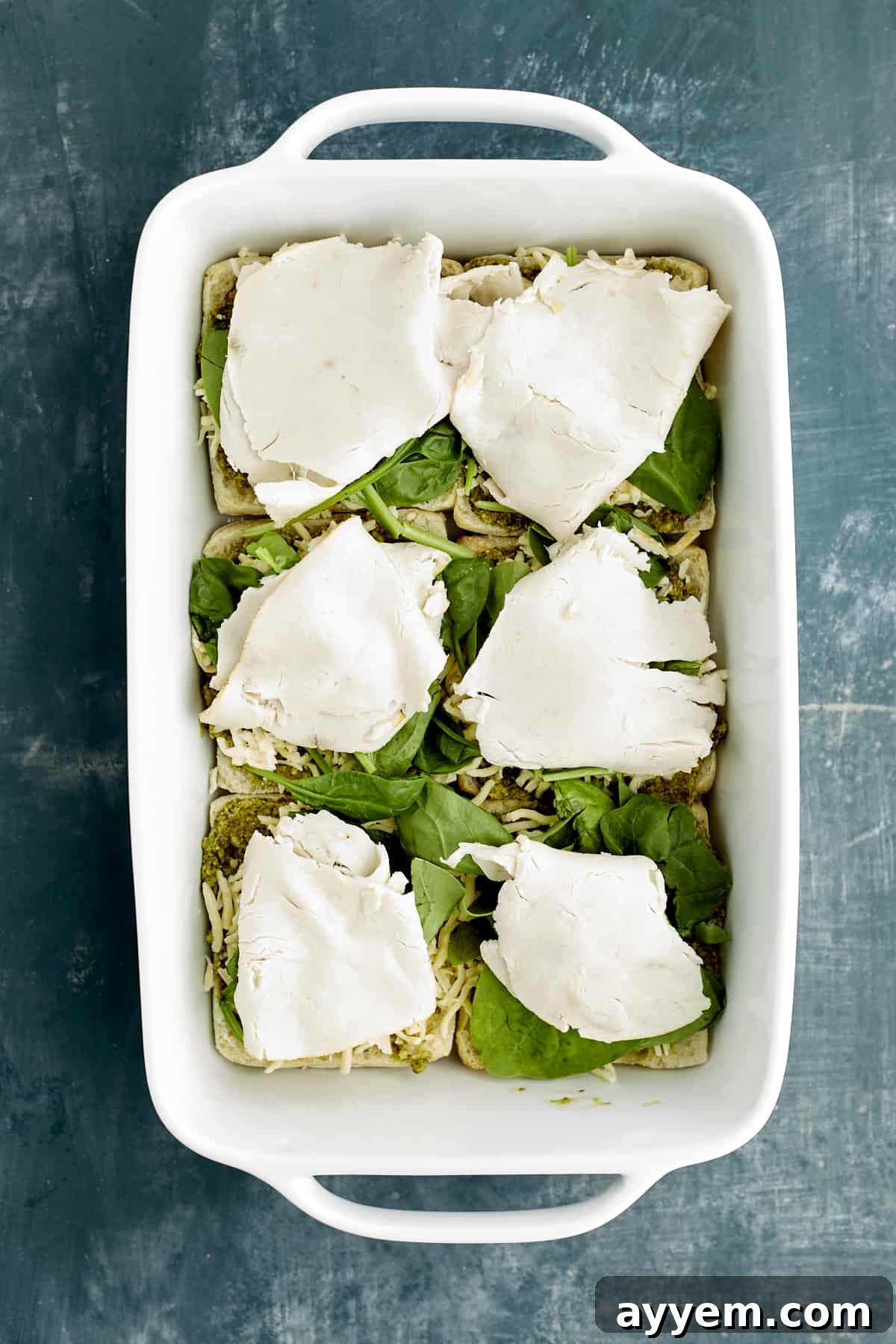 Fresh Turkey Caprese Sandwich 6 Sliced ciabatta buns in a white baking dish, layered with vibrant pesto, fresh spinach, and tender turkey slices.