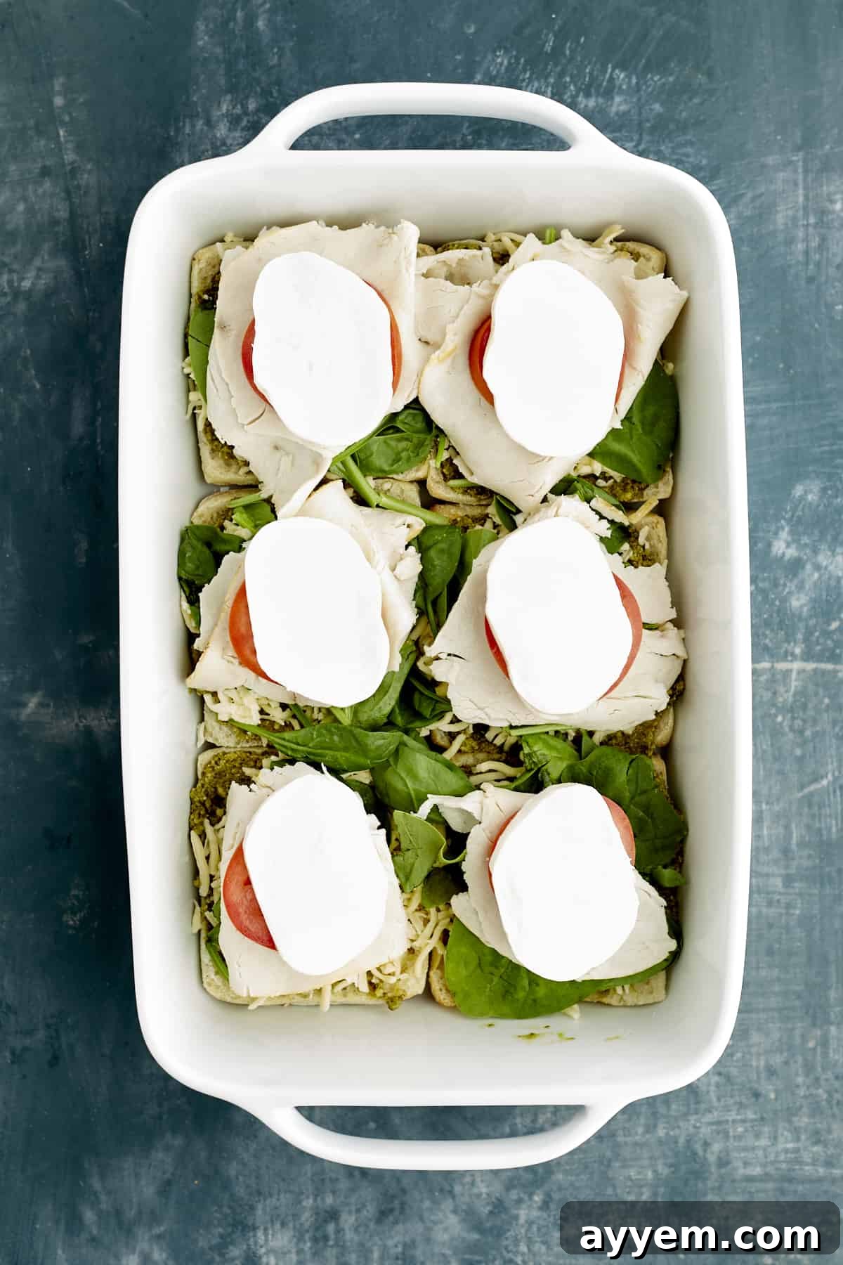 Fresh Turkey Caprese Sandwich 8 Layered ciabatta buns in a baking dish, featuring pesto, spinach, tomatoes, turkey, and generous slices of mozzarella.