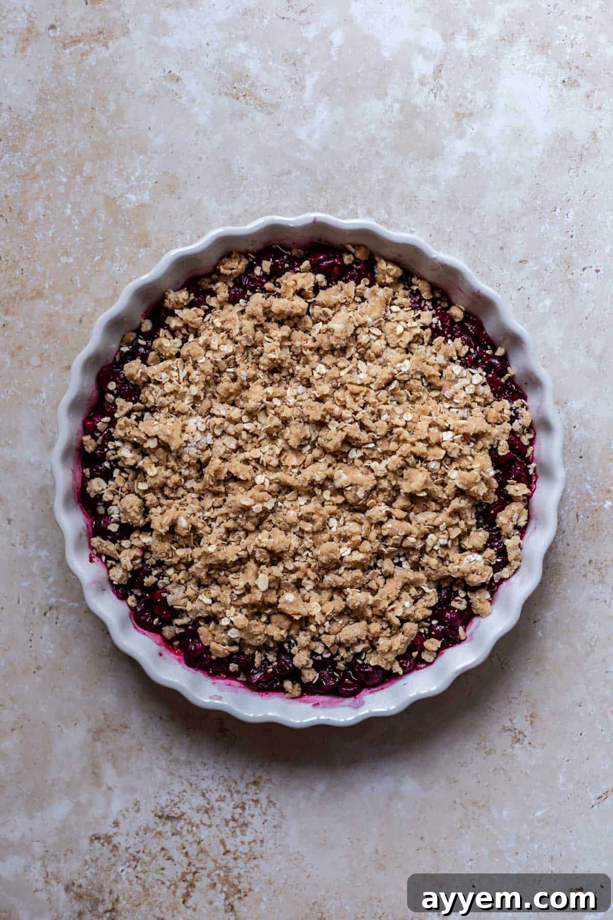An unbaked cherry crisp, featuring a smooth cherry layer beneath a generously sprinkled, golden streusel oat topping, awaiting baking.