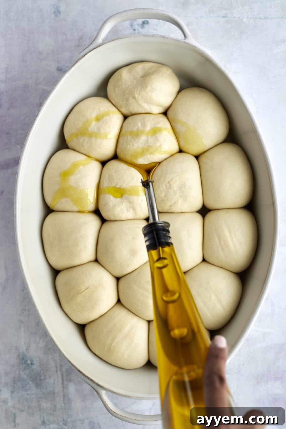 olive oil being drizzled over raw bread rolls in an oval baking dish 