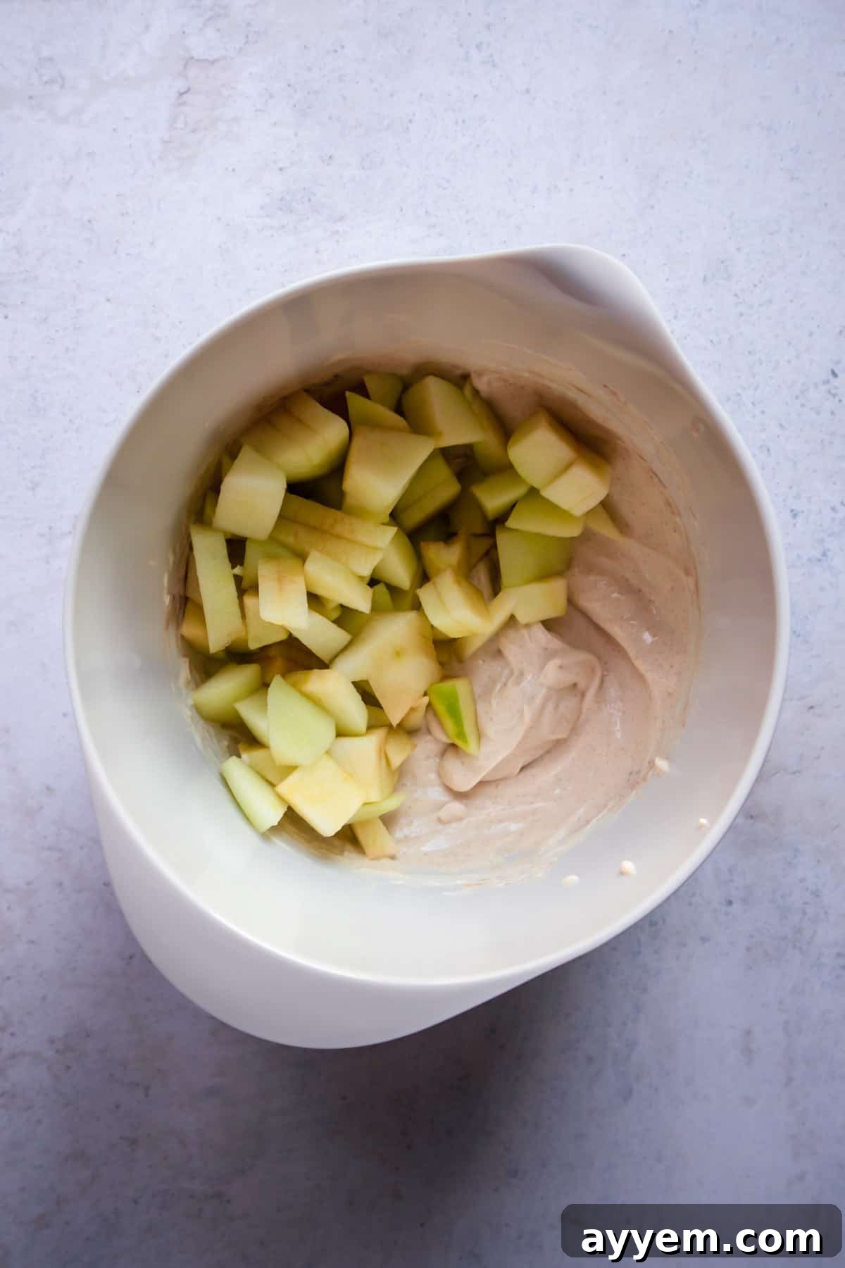 Finely chopped apples resting on top of the creamy cinnamon-cream cheese mixture in a bowl, before being folded in.