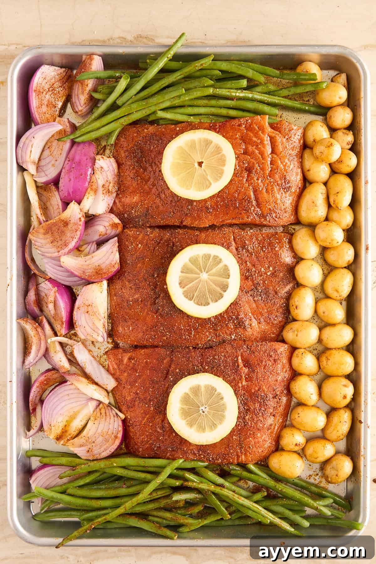 A meticulously arranged sheet pan showcasing three generously seasoned salmon fillets, each elegantly topped with a lemon slice. They are surrounded by fresh green beans, perfectly halved red onions, and small golden potatoes, all glistening and ready for their transformation in the oven.
