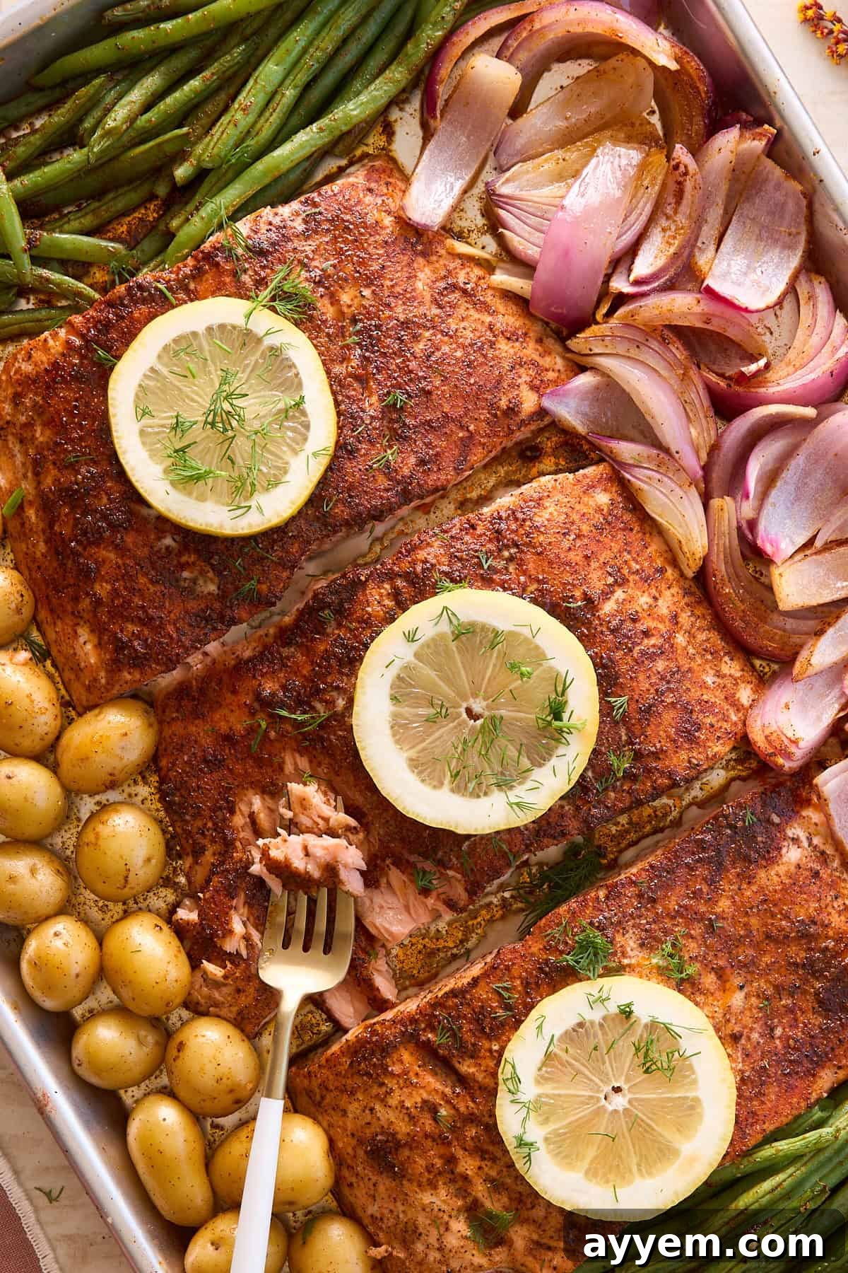 A freshly baked sheet pan featuring golden-brown salmon fillets, each adorned with a lemon slice and a sprig of fresh dill, nestled among perfectly roasted green beans, caramelized red onion wedges, and tender small yellow potatoes. A fork gently flakes a piece of the succulent salmon, highlighting its readiness.