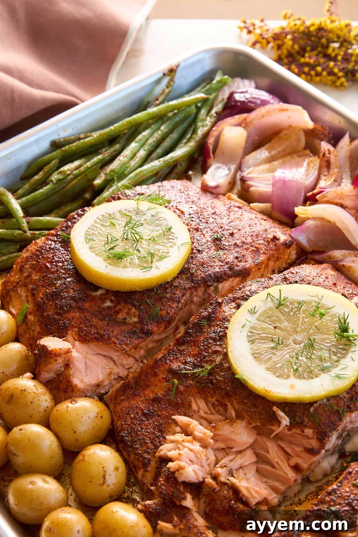 A freshly baked baking tray filled with seasoned salmon fillets, each topped with a lemon slice, alongside roasted baby potatoes, vibrant green beans, and tender red onions. The dish is garnished with fresh herbs, elegantly arranged and ready for serving.