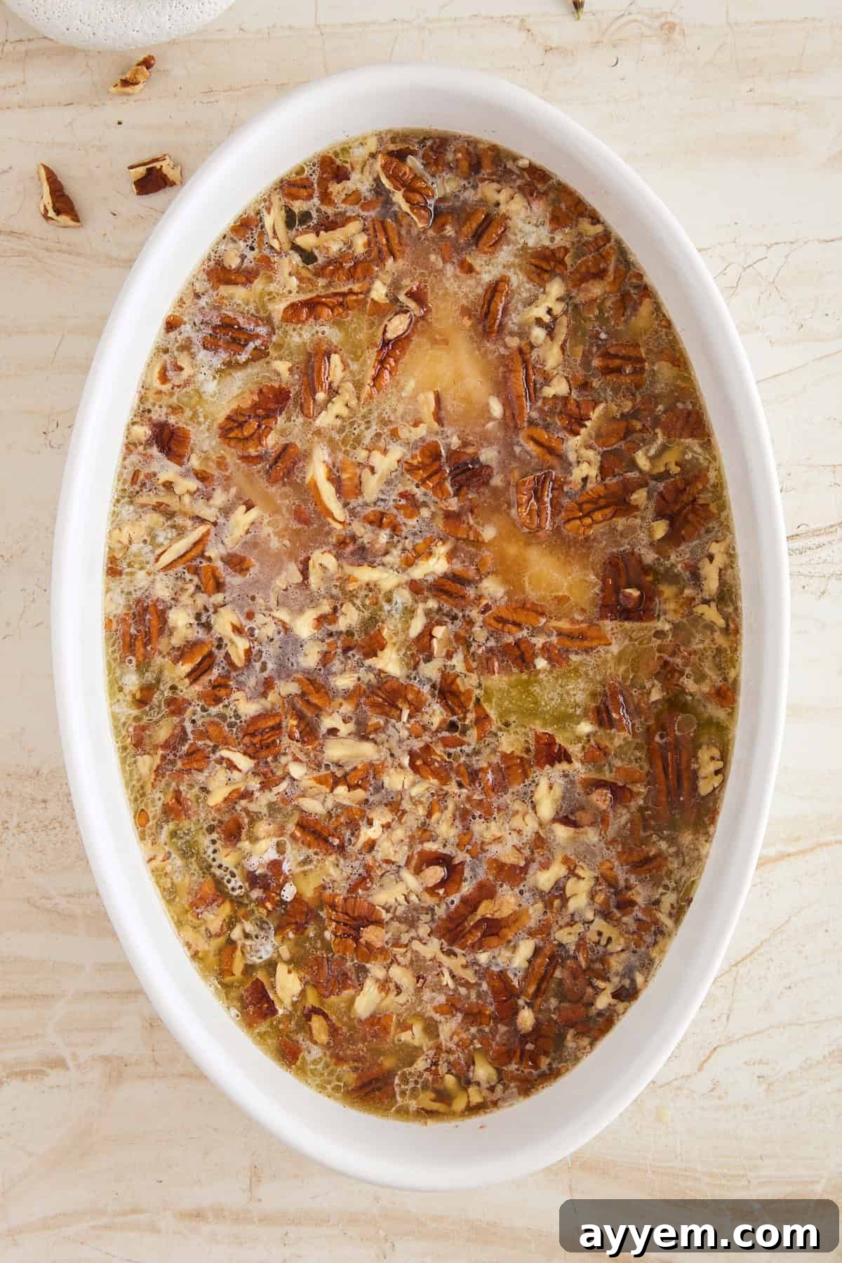 An oval white baking dish filled with a liquid mixture topped with chopped pecans, ready to be baked, sits on a light marble surface.