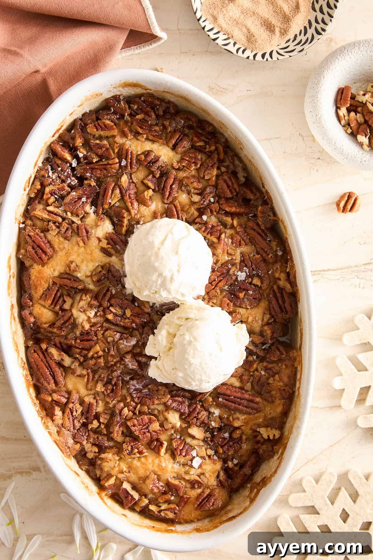 An oval dish filled with pecan bread pudding topped with two scoops of vanilla ice cream, surrounded by a napkin, a bowl of pecans, and snowflake-shaped decorations on a beige surface.