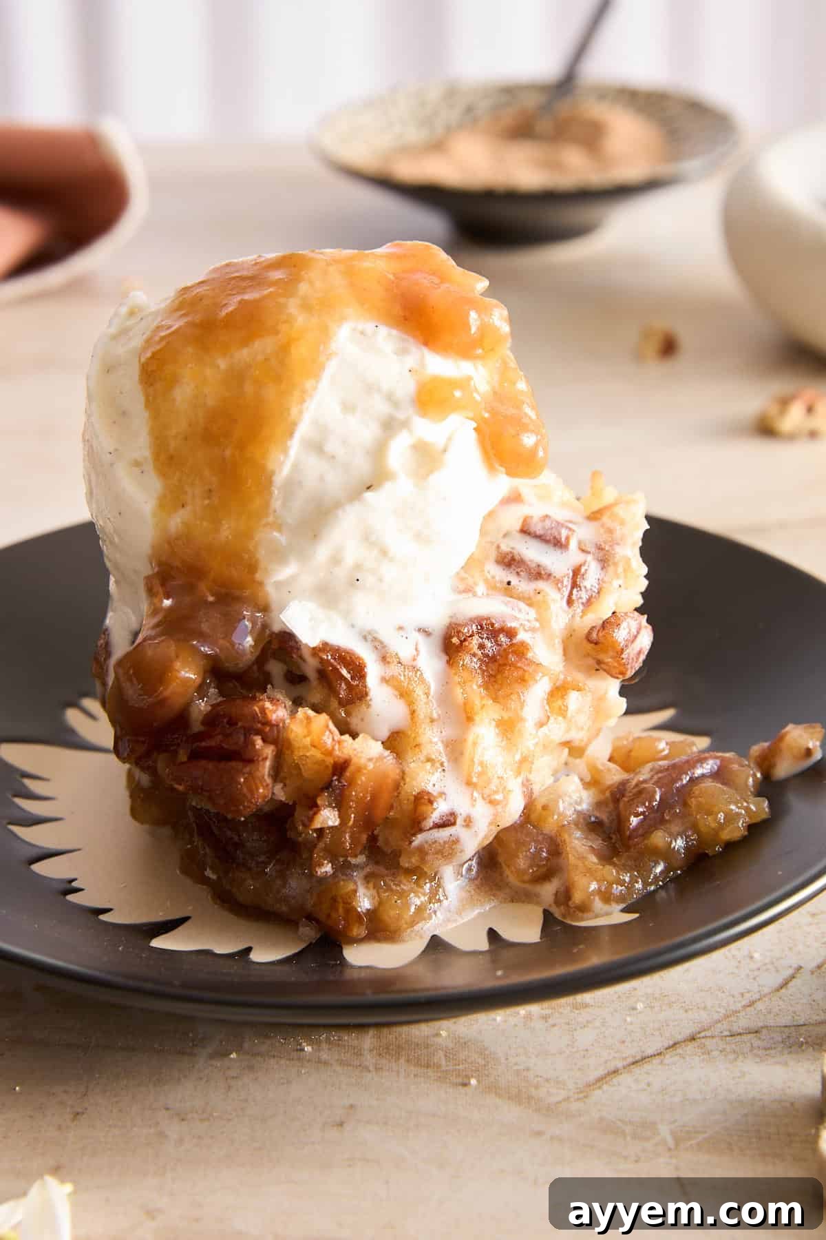 A slice of pecan cobbler topped with a scoop of vanilla ice cream and caramel sauce sits on a black plate, with melted ice cream dripping down the sides. A bowl and spoon are blurred in the background.