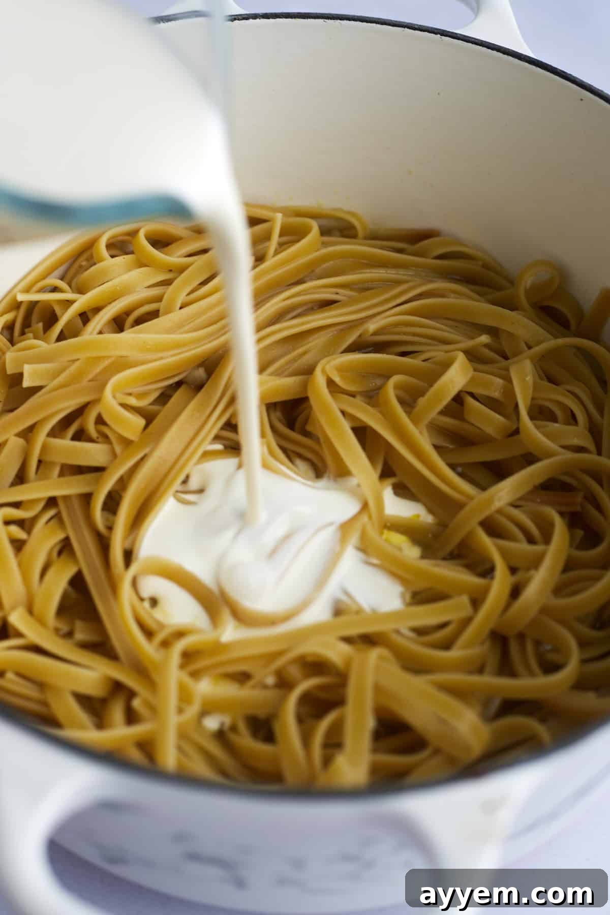 Luxurious alfredo sauce being poured over perfectly cooked fettuccine noodles directly in the cooking pot, ready to be combined.