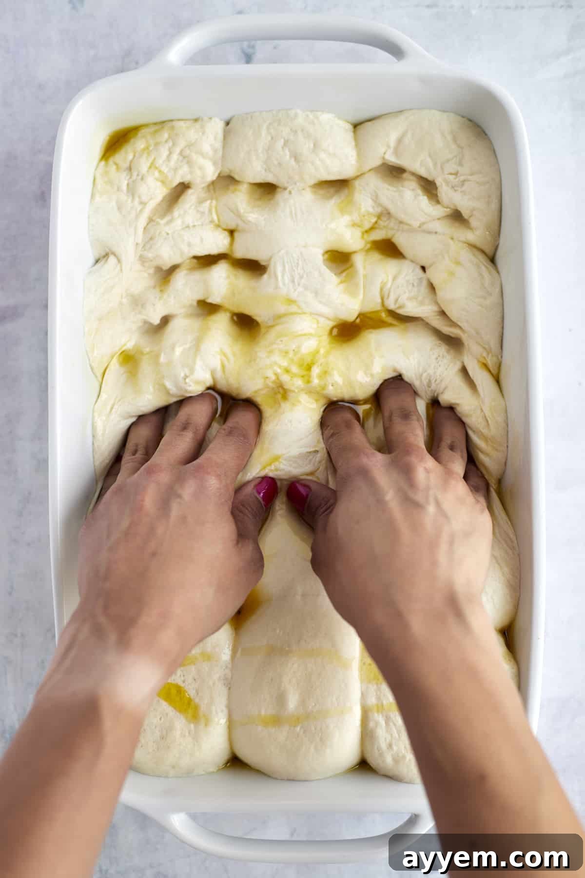 Fingers pressing characteristic dimples into olive-oil-drizzled focaccia dough.