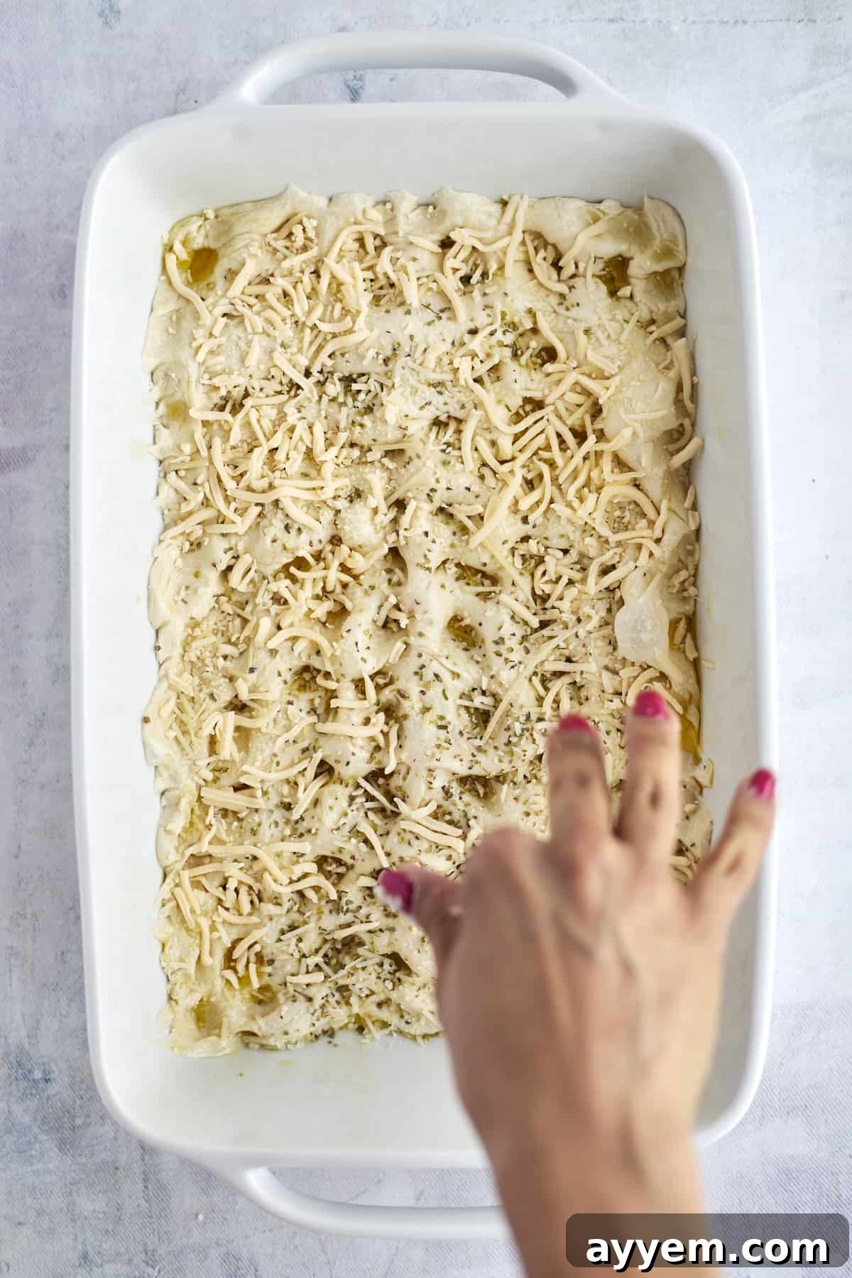 Shredded cheese being sprinkled generously over unbaked focaccia bread rolls.