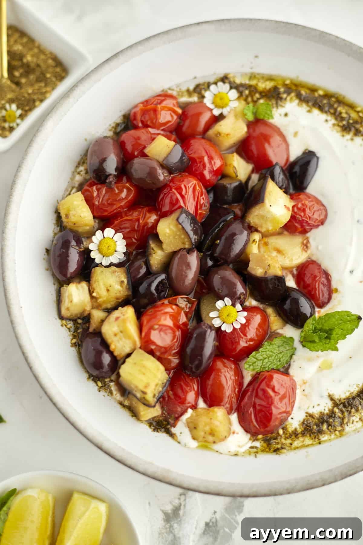 Plated eggplant, tomatoes and olives over creamy yogurt dip. Garnished with zaatar and fresh mint
