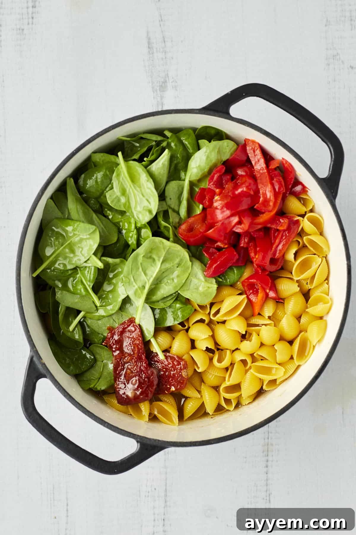 Cooked pasta shells, vibrant red bell peppers, and fresh spinach tossed in a large pot, forming the base of the Tuscan chicken pasta.