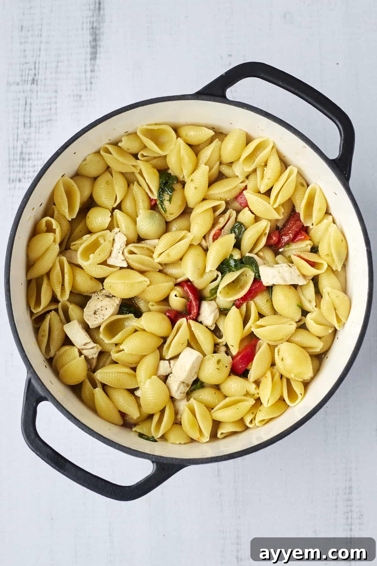 A vibrant mix of cooked pasta shells, fresh spinach, roasted red peppers, and chicken in a large pot, ready for the final creamy touch.