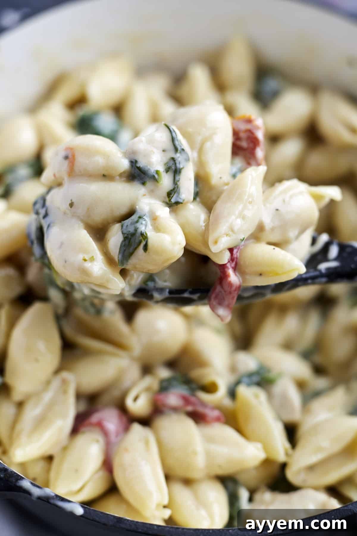 A generous spoonful of creamy Tuscan chicken pasta being lifted from a large pot, showcasing the rich sauce and perfectly cooked ingredients.