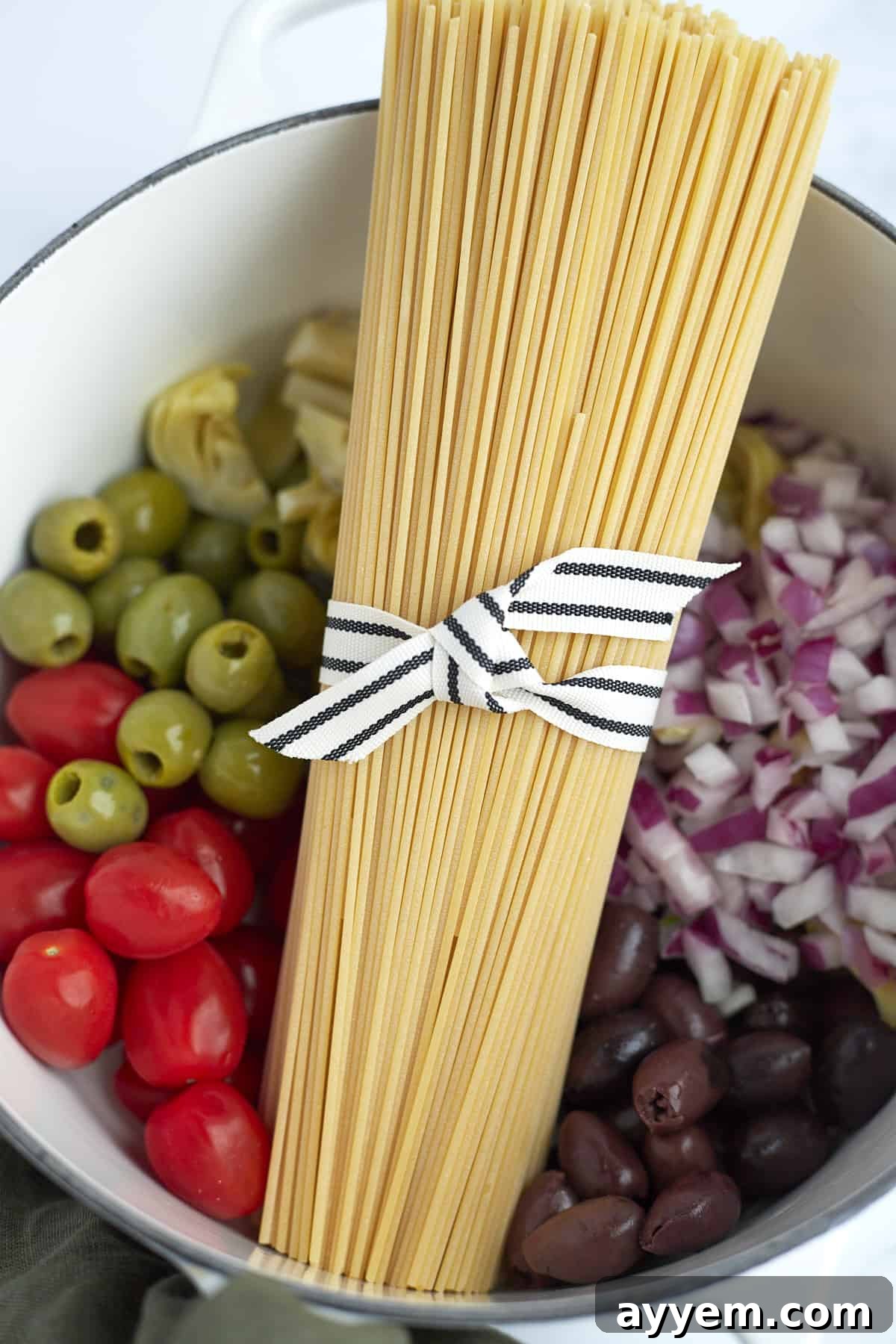 An overhead view inside a pot filled with green and black olives, bright cherry tomatoes, finely chopped red onion, and a bundle of uncooked spaghetti noodles, all prepped for cooking.