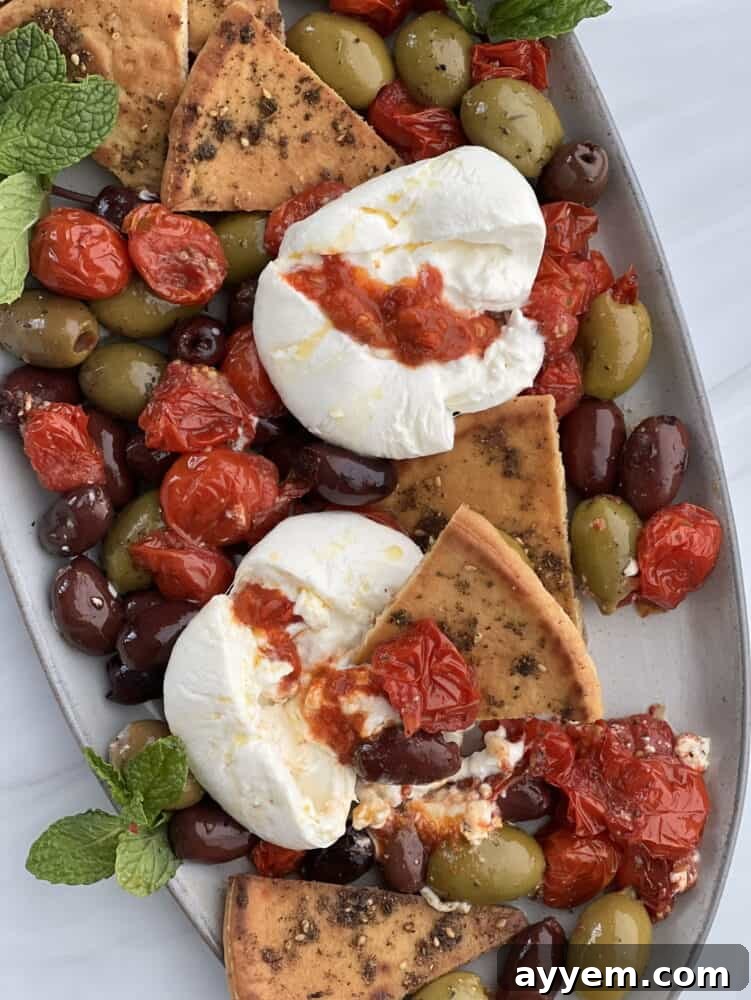 close up of burrata with a piece missing, showing creamy interior, surrounded by roasted tomatoes, olives and zaatar pita