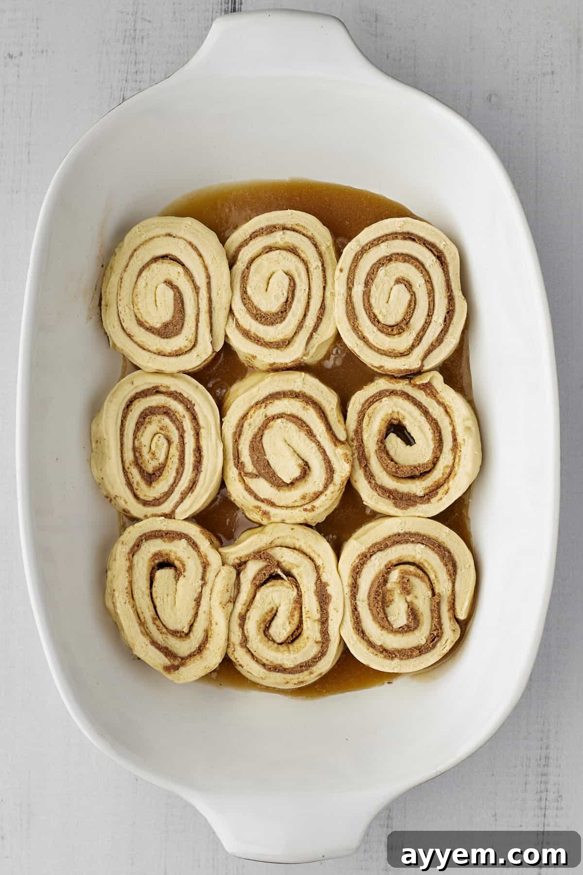 Unbaked cinnamon rolls carefully arranged on a bed of homemade caramel sauce in a white baking dish, showing the preparation stage.