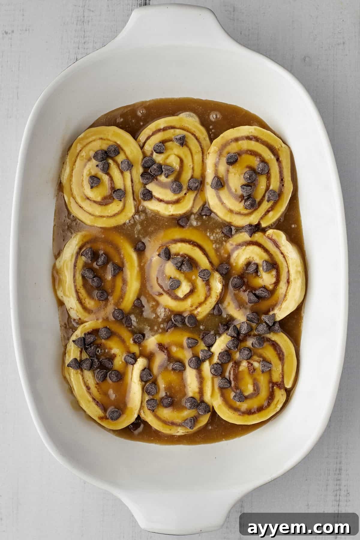A white baking dish filled with unbaked cinnamon rolls, coated in caramel sauce and generously topped with chocolate chips, anticipating baking.