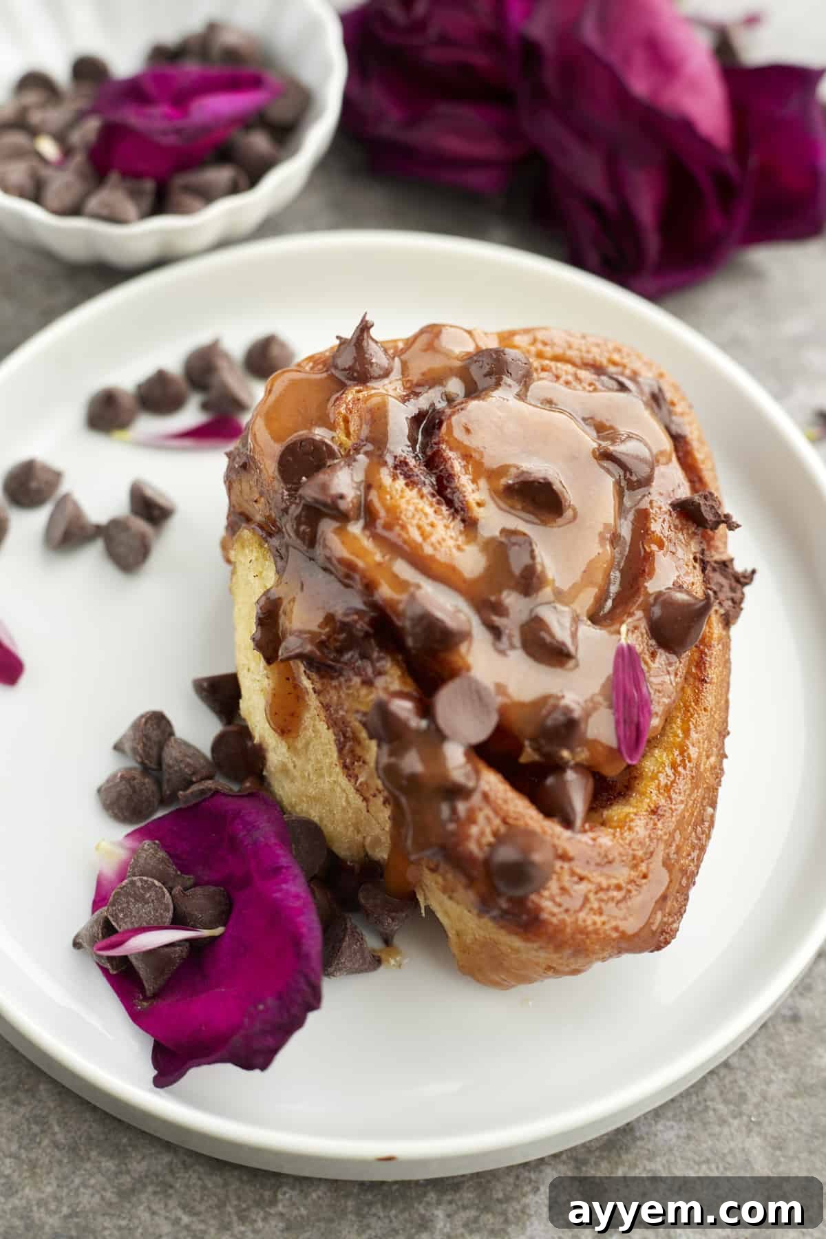 A single, perfectly golden caramel chocolate chip cinnamon roll on a white plate, garnished with scattered chocolate chips and a delicate pink rose, highlighting its gourmet appeal.