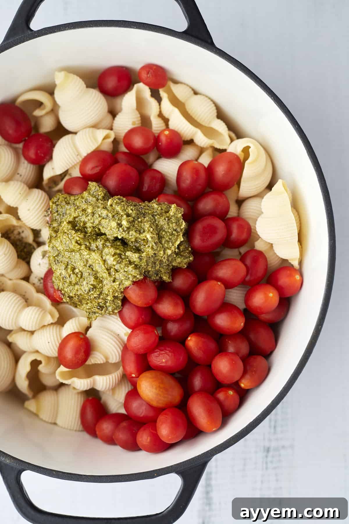 Large pot filled with jumbo pasta shells, vibrant cherry tomatoes, and fresh pesto before cooking.