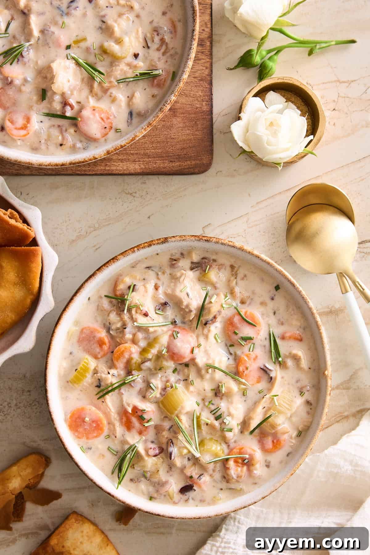 A beautifully presented bowl of creamy chicken wild rice soup, garnished with fresh herbs, sits on a rustic wooden table. A gold spoon rests beside the bowl, with toasted bread and a smaller bowl of a complementary dish in the background, creating an inviting and warm scene.