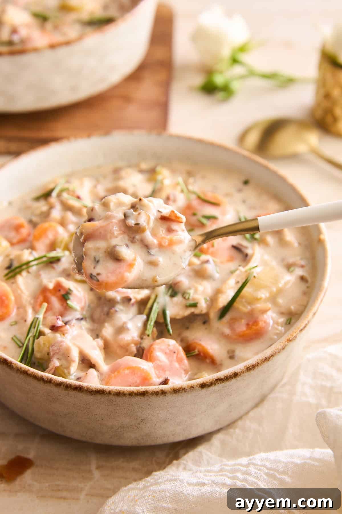 A close-up shot of a rustic bowl of creamy chicken wild rice soup, showing visible chunks of tender chicken, vibrant carrots, earthy mushrooms (if added), and fresh herbs. A golden spoon is positioned, ready to lift a generous portion of the hearty soup, with a soft cloth napkin and blurred background suggesting a warm dining setting.