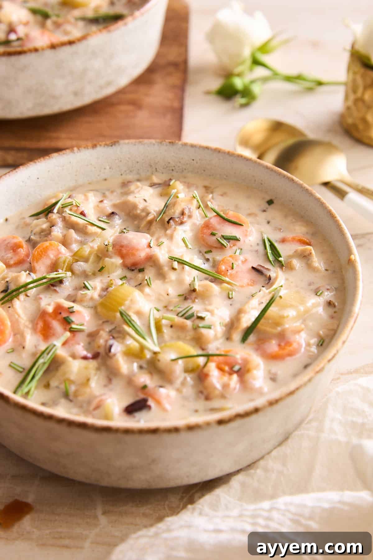 A inviting bowl of creamy chicken wild rice soup, generously filled with tender chicken pieces, crisp carrots, fresh celery, and hearty wild rice. It's elegantly garnished with fresh rosemary and chives, placed on a bright table alongside gleaming gold spoons. A delicate white flower and a blurred second bowl in the background complete this comforting culinary tableau.
