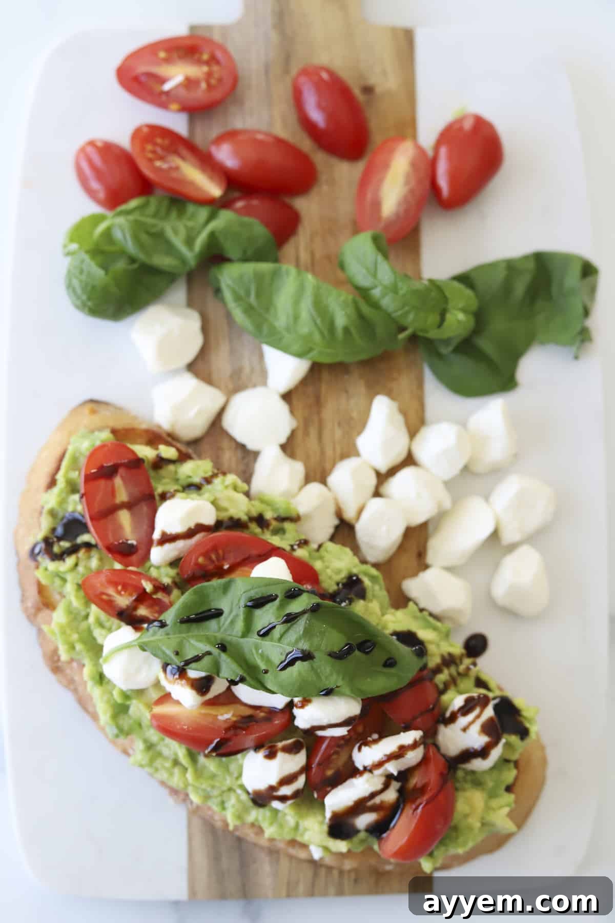 A close-up overhead shot of a finished Caprese Avocado Toast, showing the rich green avocado spread, glistening red tomatoes, white mozzarella pearls, dark balsamic glaze, and fresh green basil on toasted sourdough.